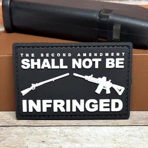 May include: Black rectangular patch with white text and gun illustrations. The text reads "THE SECOND AMENDMENT SHALL NOT BE INFRINGED." A rifle and a modern sporting rifle are depicted. A black object is in the background.