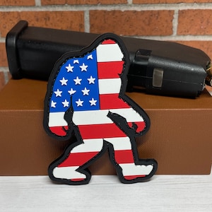 May include: A red, white, and blue American flag design on a black patch shaped like a Bigfoot walking.