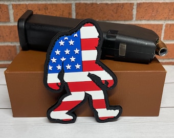 May include: A red, white and blue American flag design on a black patch shaped like a Bigfoot walking.