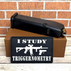 May include: Black firearm magazine with a single bullet visible, resting on a brown box. A black patch below reads "I STUDY TRIGGERNOMERTY" with white text and gun illustrations. The background features a brick wall.