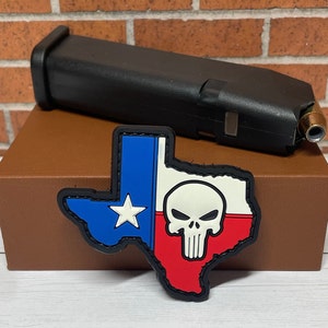 May include: A red, white, and blue Texas-shaped patch with a white skull and crossbones in the center. The patch is on a brown box.