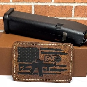 May include: A brown leather patch with a black and white American flag design. The flag has a silhouette of a rifle and the text "F.A.T.O." with a target symbol.