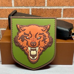 May include: A shield-shaped patch featuring a snarling wolf's head in shades of orange, brown, and white, set against a green background. The patch has a brown border and is displayed on a brown box.