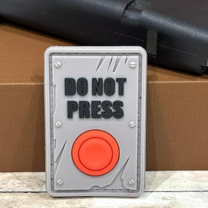 May include: A gray and red patch with the text "DO NOT PRESS" and a red button in the center. The patch is made of a textured material and has a distressed look.