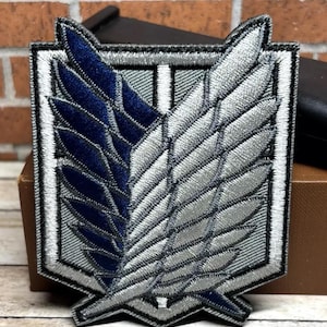 May include: A gray and blue embroidered patch with a winged symbol, resembling a stylized bird, inside a gray rectangle with a white border. The patch is a popular symbol from the anime series Attack on Titan.