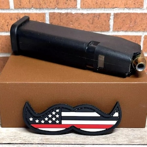 May include: A black magazine for a handgun with a red, white, and blue thin red line American flag patch in the shape of a mustache.