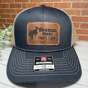 May include: A navy blue and tan trucker hat with a brown leather patch that says "Bronco Hair Don't Care". The hat is adjustable and has a Richardson logo on the back.