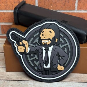 May include: A black and gray embroidered patch featuring a cartoon character wearing a suit and holding a gun. The character has a black beard and a black and white patterned background.