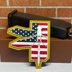 May include: A yellow embroidered patch with a white dragonfly silhouette on a red, white, and blue American flag background.
