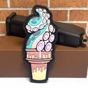 May include: A colorful patch featuring an ice cream cone design with an octopus tentacle. The ice cream is pink, with blue and green tentacles, and a yellow cone. The patch has a black border.