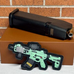 May include: A green and black tactical rifle patch with a black magazine and a red dot sight. The patch is on a brown box with a black magazine on top.