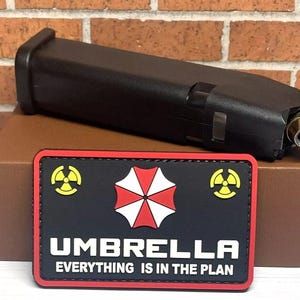 May include: Black and red embroidered patch with a white umbrella logo and the text "Umbrella Everything is in the Plan" with yellow biohazard symbols on either side.