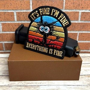 Peut inclure: Un patch brod&eacute; noir avec un fond de coucher de soleil vintage d&eacute;lav&eacute;. Le patch repr&eacute;sente un monstre de dessin anim&eacute; avec de grands yeux et le texte "It's Fine I'm Fine Everything Is Fine".