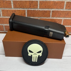May include: A black plastic magazine for a handgun with a glow-in-the-dark skull patch. The magazine is sitting on a brown box.