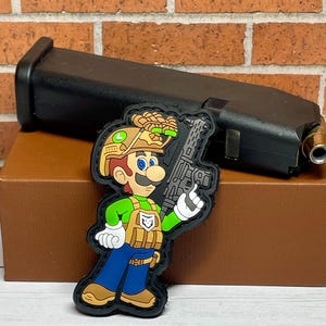 May include: A green and brown cartoon character wearing a helmet and holding a gun. The character is wearing a tactical vest and blue jeans. The character is a parody of the video game character Mario.