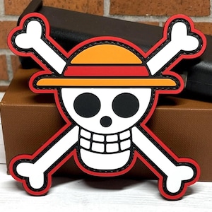 May include: A colorful patch featuring the Jolly Roger symbol from the One Piece anime. The design includes a white skull with black eyes, a straw hat, and crossed bones, outlined in red and black.