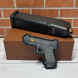 May include: A black and gray handgun patch with a yellow accent. The patch is on a brown box with a black magazine on top.