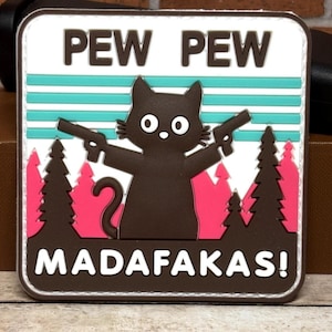 May include: A square patch featuring a cartoon cat holding two guns. The patch has the words "PEW PEW" at the top and "MADAF*KAS!" at the bottom. The cat is brown, the background is white with teal stripes, and pink trees.