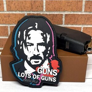 May include: Black and white patch with a stylized image of a man's face. The patch has pink, blue, and red accents and the text "GUNS LOTS OF GUNS PRINCIPLE".