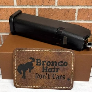 May include: A brown leather patch with the text "Bronco Hair Don't Care" and a silhouette of a horse's head. The patch is sitting on top of a brown box with a black gun magazine on top of the box.