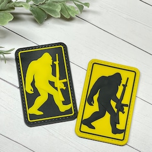 May include: Two yellow and black embroidered patches featuring a silhouette of Bigfoot holding a rifle. The patches are rectangular and have a black border.