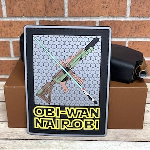 May include: A rectangular patch with a black border features a stylized weapon design, a light blue lightsaber, and the text "OBI-WAN NAIROBI" in yellow and white. The background has a gray honeycomb pattern.