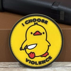 May include: A round yellow patch with a cartoon duck holding a knife. The patch has black text that reads "I CHOOSE VIOLENCE" around the duck. The duck has a red beak and feet. The patch is surrounded by a black border.