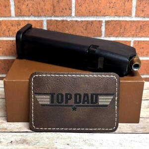May include: Brown leather patch with white stitching and the text "TOP DAD" in a white font with a white star underneath. The text is in a stylized font similar to the "Top Gun" logo.