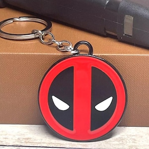 May include: A red and black metal keychain featuring the Deadpool logo. The logo is a circle with a red center and a black outline. The center of the circle has a black vertical line with two white eyes on either side of the line.