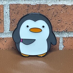 May include: A black and white enamel pin of a penguin holding a knife. The penguin has a black head and beak, white body, and orange beak. The knife is black with a pink handle.