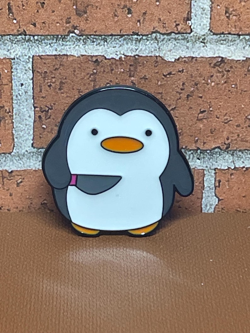 May include: A black and white enamel pin of a penguin holding a knife. The penguin has a black head and beak, white body, and orange beak. The knife is black with a pink handle.
