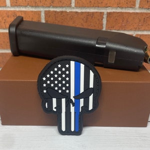 May include: A black and blue skull patch with an American flag and a thin blue line. The patch is made of fabric and has a velcro backing.