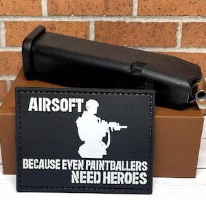 May include: Black and white patch with the text "Airsoft: Because even paintballers need heroes". A white silhouette of a person holding a gun is in the center of the patch. The patch is on top of a brown box. A black gun magazine is on top of the box.