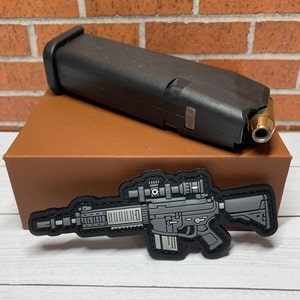 May include: A black and gray tactical rifle patch with a scope and a silencer. The patch is on a brown box with a black magazine on top.