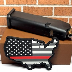 May include: A black magazine for a handgun with a red, white, and gray American flag patch with a thin red line. The patch is shaped like the United States.