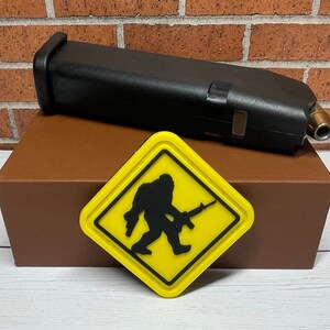 May include: A yellow warning sign with a black silhouette of a Bigfoot holding a rifle. The sign is on a brown box with a black gun magazine on top.