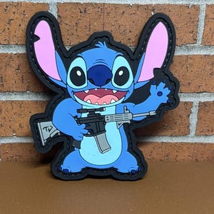 May include: A blue cartoon character patch with pink ears and a black outline. The character is holding a black and silver rifle. The patch is on a brown surface with a brick wall background.