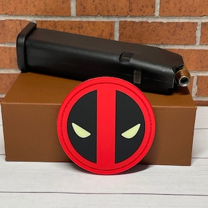 May include: A brown cardboard box with a red and black Deadpool logo patch on top. The box is sitting on a white surface.