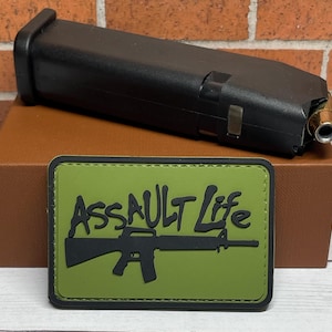 May include: A black firearm magazine with a brass-colored bullet is positioned above a green patch. The patch features the words "ASSAULT LIFE" in black, along with a black rifle silhouette. The items rest on a brown box, against a brick background.