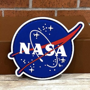 May include: A blue, white, and red NASA logo patch. The patch features the NASA logo with the word "NASA" in white, a red swoosh, and white stars on a blue background. The patch is circular with a white border.