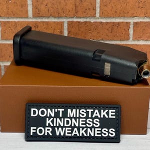 May include: A black magazine for a handgun with a brass bullet in the magazine well. The magazine is sitting on a brown box with a black patch that reads "Don't mistake kindness for weakness".