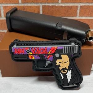 May include: A black and gray pistol-shaped patch with the text "BABA YAGA" in red, orange, and purple. The patch features a stylized image of a man in a suit. A black magazine and a bullet are in the background.