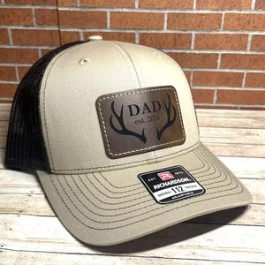 May include: A beige and black trucker hat with a brown leather patch that says "DAD est. 2024" with a pair of antlers. The hat is adjustable and has a black mesh back.