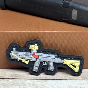 May include: A gray and yellow patch in the shape of a rifle with a black border. The rifle has a red accent and a yellow sight. The patch is on a brown surface with a black object in the background.