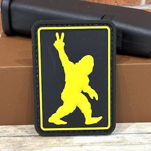 May include: A black and yellow patch with a silhouette of a Bigfoot figure giving the peace sign.