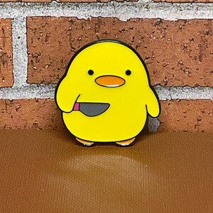 May include: Yellow enamel pin of a cartoon chick holding a knife. The chick has black eyes and a pink beak.