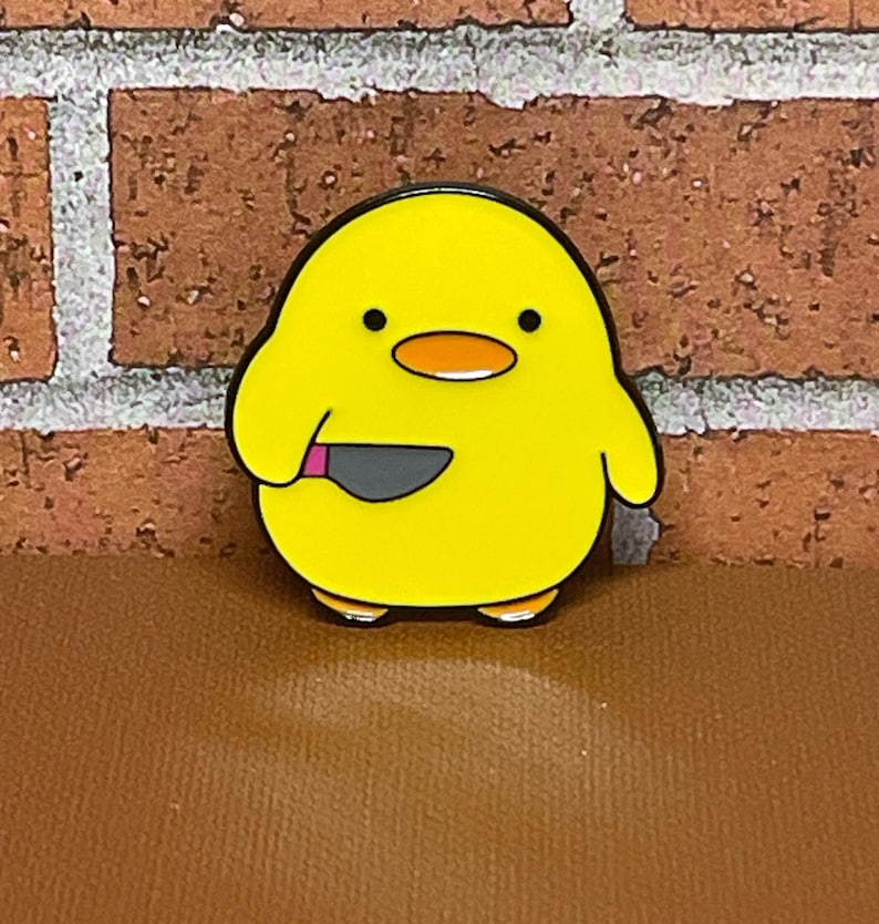 May include: Yellow enamel pin of a cartoon chick holding a knife. The chick has black eyes and a pink beak.