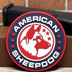 May include: A round patch with the words "AMERICAN SHEEPDOG" in white lettering. The patch features a red and white dog silhouette, surrounded by a blue ring with red stars. The patch is on a brown surface with a black object in the background.