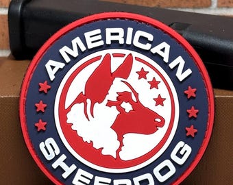 May include: A round patch with the words "AMERICAN SHEEPDOG" in white lettering. The patch features a red and white dog silhouette, surrounded by a blue ring with red stars. The patch is on a brown surface with a black object in the background.