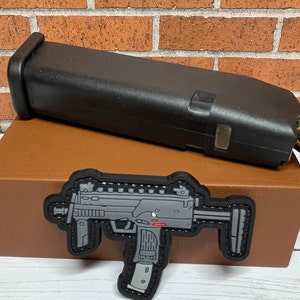 May include: A black plastic magazine for a firearm, a brown box, and a gray and black patch with a stylized image of a submachine gun.